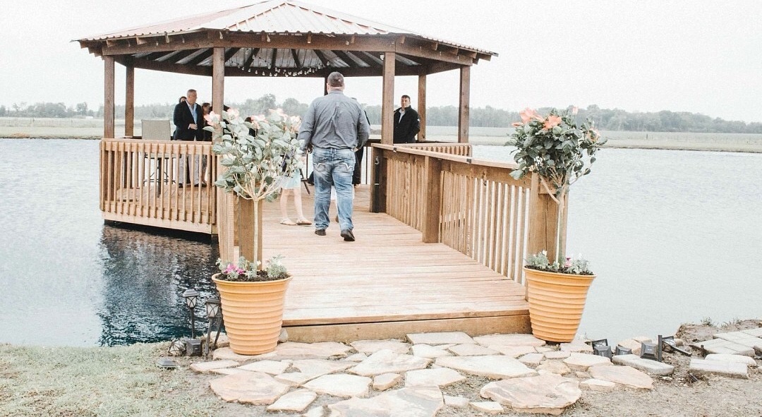 Wooden gazebo over the water with floral arrangements flanking a stone path — a warm welcome venue for ceremonies